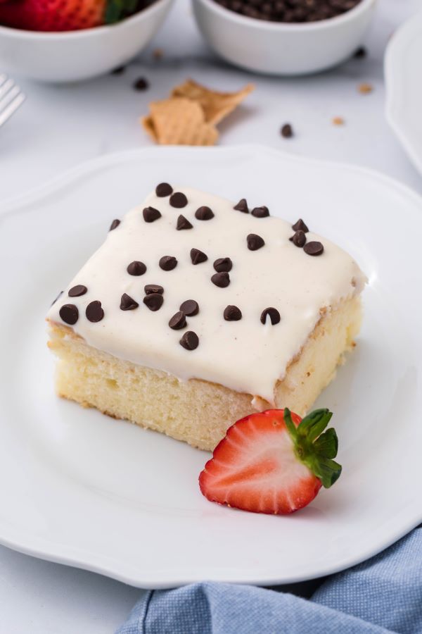 Slice of Cannoli Poke cake on a white plate and a strawberry for garnish,.