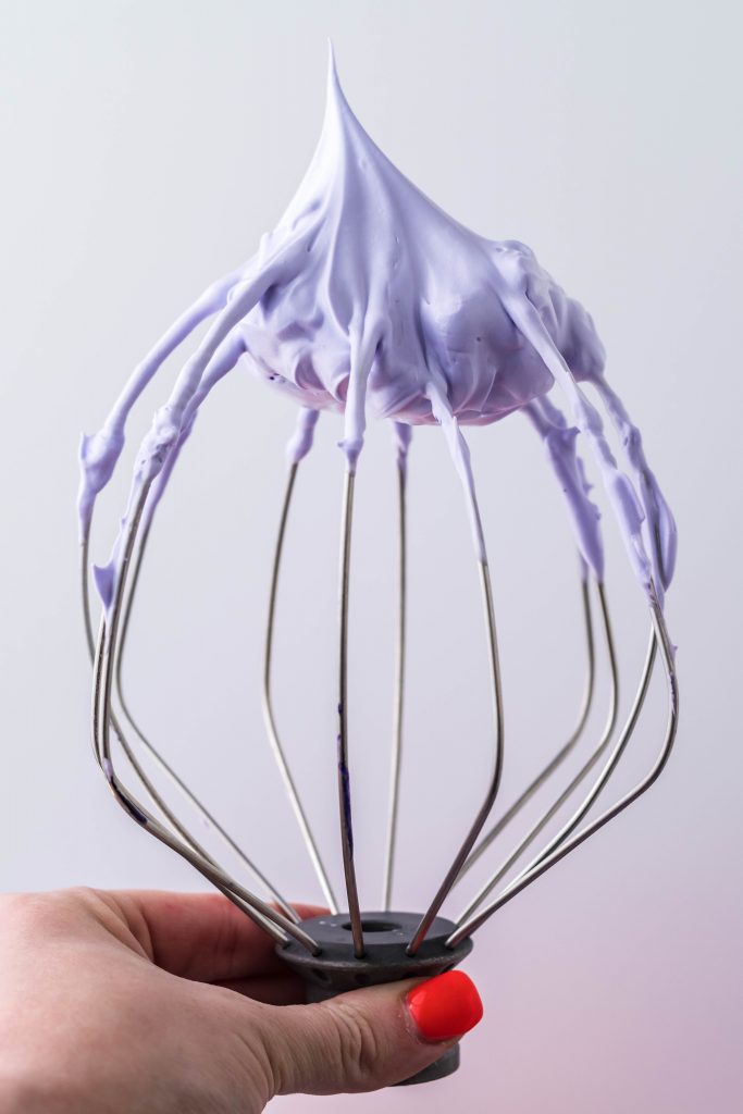 A stand mixer whisk attachment with lavender colored stiff peak on top