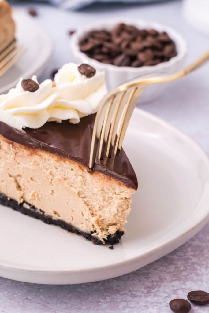 A slice of mocha cheesecake on a white plate with a gold fork removing a bite. 