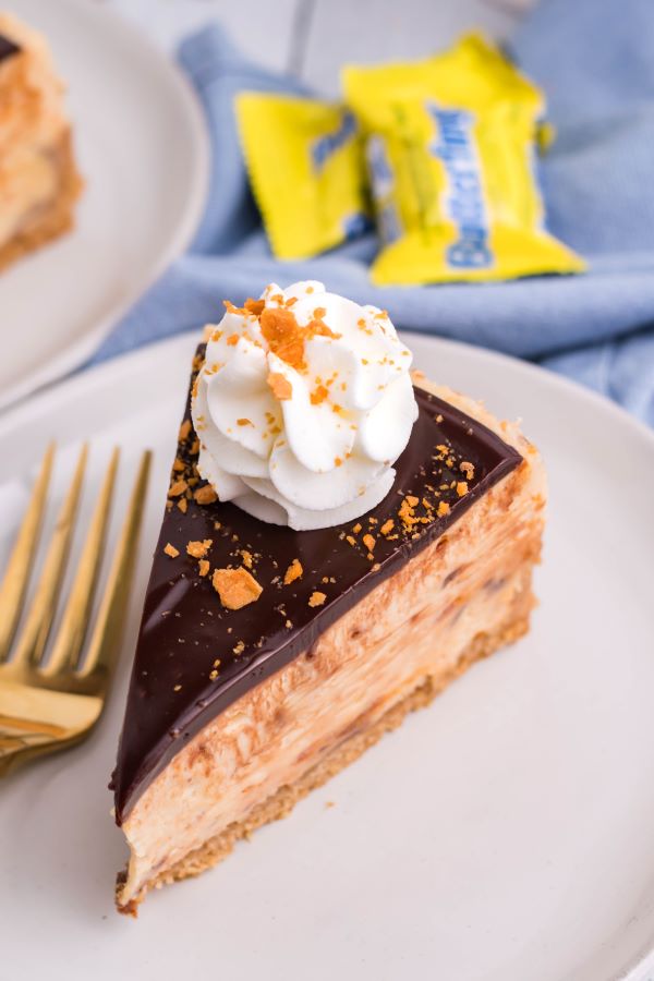 A slice of Butterfinger cheesecake on a white plate with a  gold fork.
