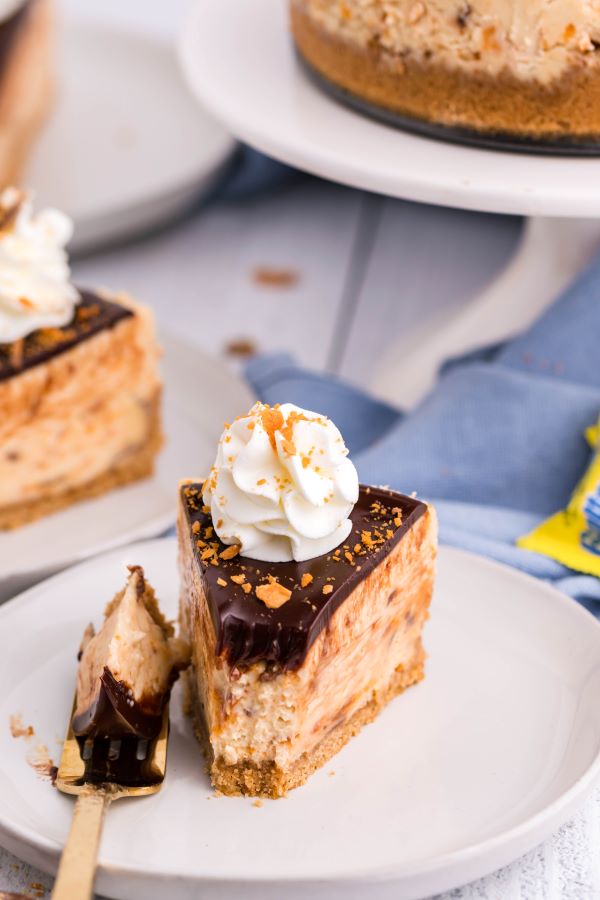 A slice of Butterfinger cheesecake on a white plate with a blue linen and crushed butterfingers in the background.