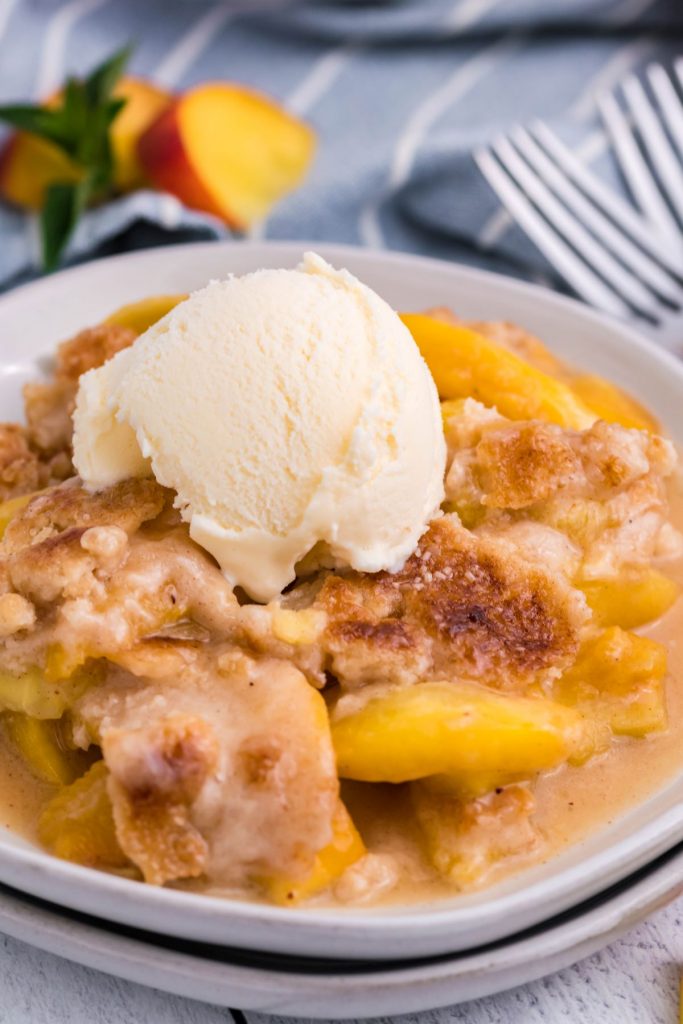 Instant Pot Peach Cobbler on a white plate topped with vanilla ice cream with peaches and a blue and white striped linen in the background. 