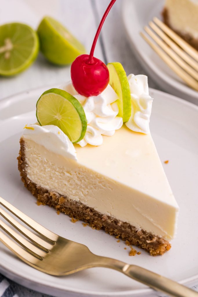 A slice of cheesecake on a white plate with a gold fork with limes in the background.