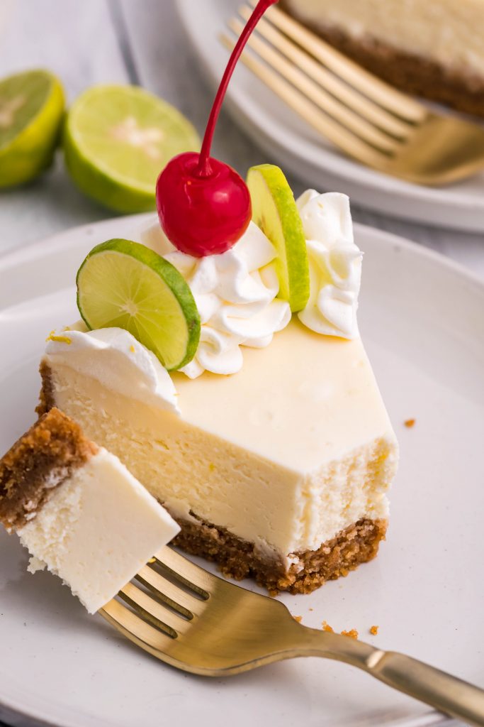 A slice of cheesecake on a white plate with a bite removed onto a gold fork with limes in the background.