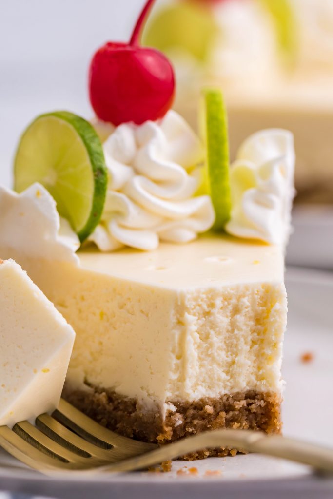 Up close view of a slice of cheesecake on a white plate with a bite removed onto a gold fork with limes in the background.