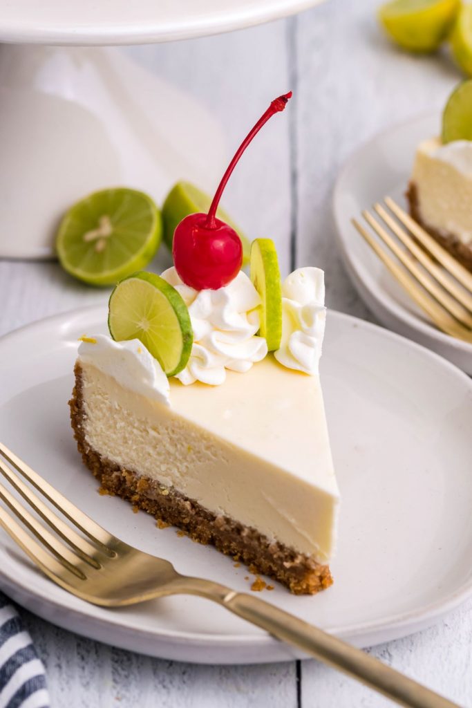 A slice of cheesecake on a white plate with a gold fork with limes in the background. 