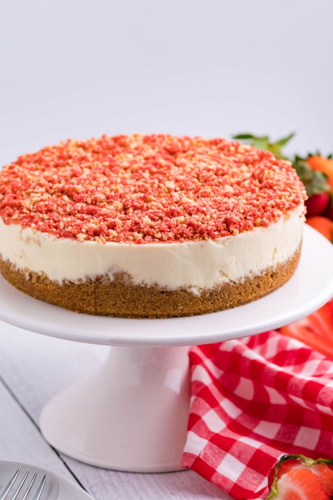 The full cheesecake on a white cake stand with a red and white checkered linen and strawberries in the background.