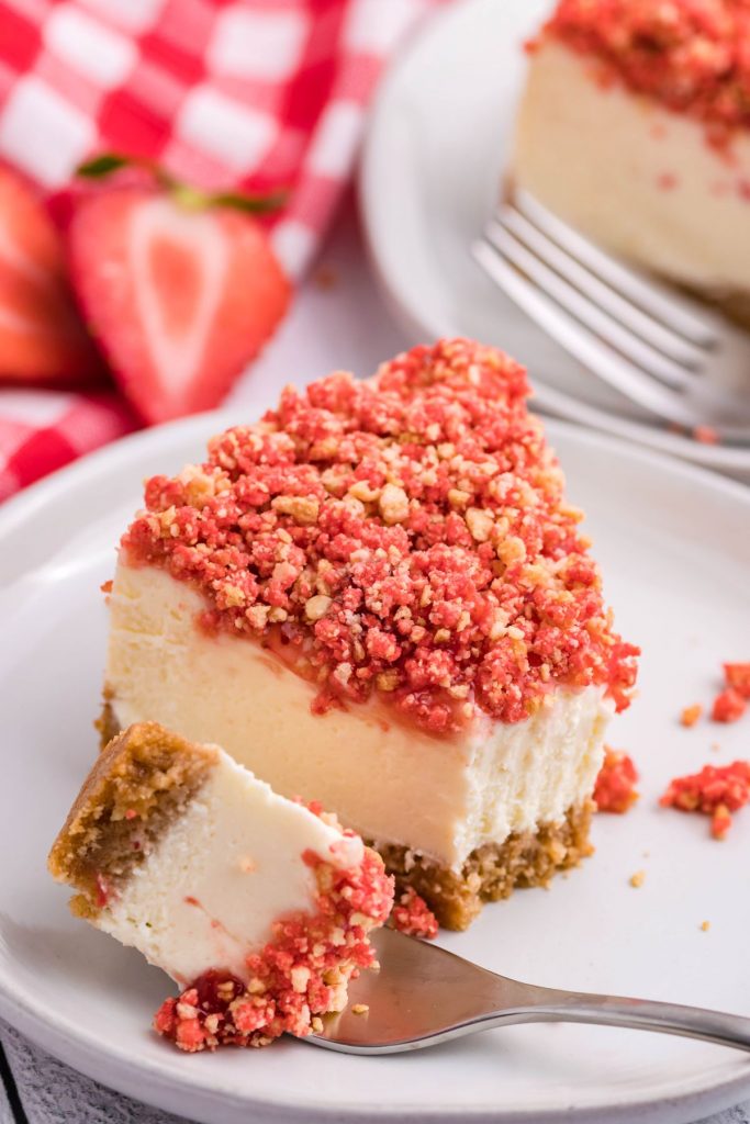 A piece of strawberry crunch cheesecake on a white plate with a bite removed onto a silver fork. 
