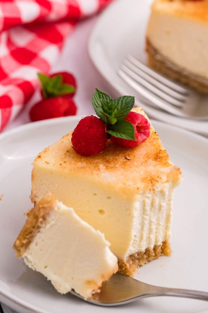 A piece of Instant Pot Crème Brulee Cheesecake on a white plate with a bite removed. Topped with raspberries and fresh mint. 