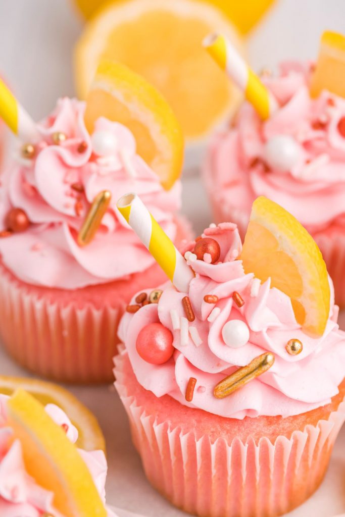 45 degree  view of pink lemonade cupcakes with sprinkles, a lemon slice, and a white and yellow straw.