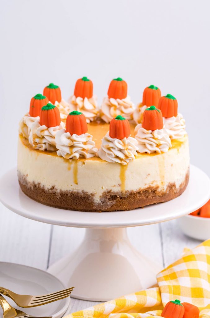The full cheesecake on a white cake stand with a yellow and white checkered linen in the background. Topped with pumpkin candy and caramel drizzle.