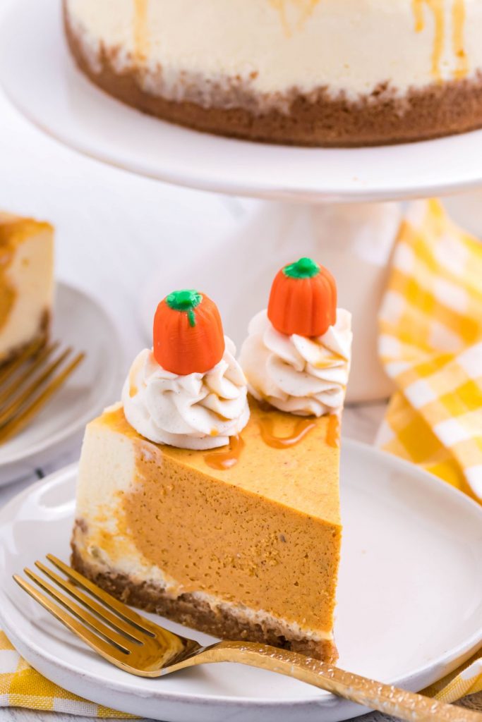 A slice of Instant Pot Pumpkin Cheesecake on a white plate with a gold fork. Topped with pumpkin candy and caramel drizzle.