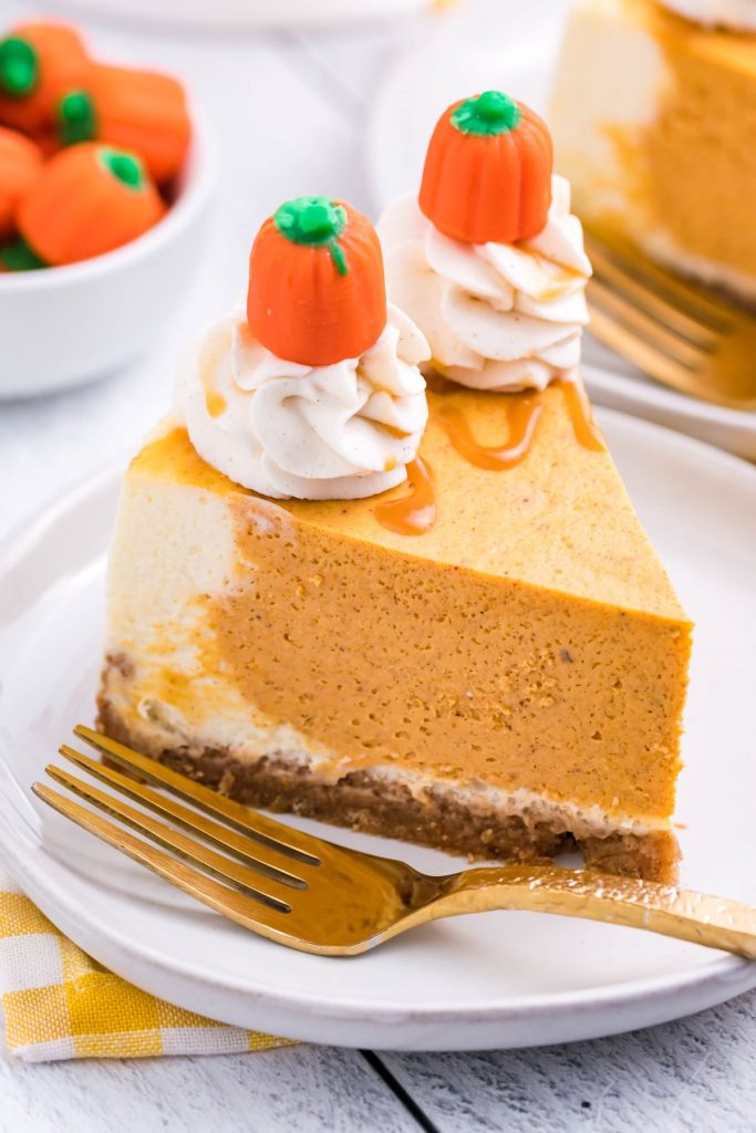 A slice of Instant Pot Pumpkin Cheesecake on a white plate with a gold fork. with a yellow and white linen and pumpkin candy in the background.