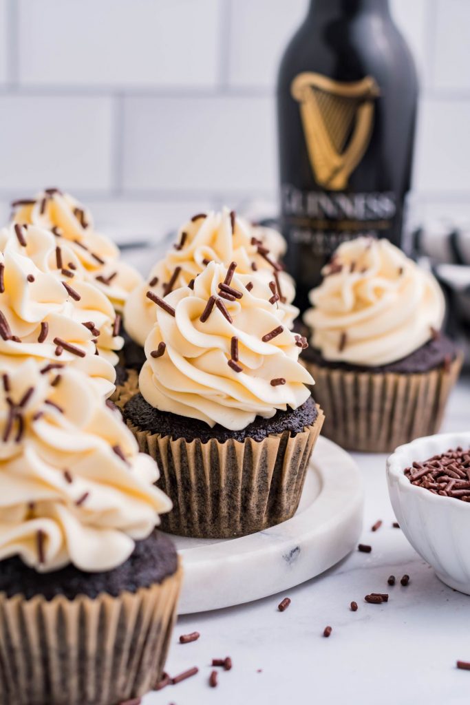 Side view of Guinness Cupcakes with white buttercream frosting and brown sprinkles. With a Guinness bottle in the background.