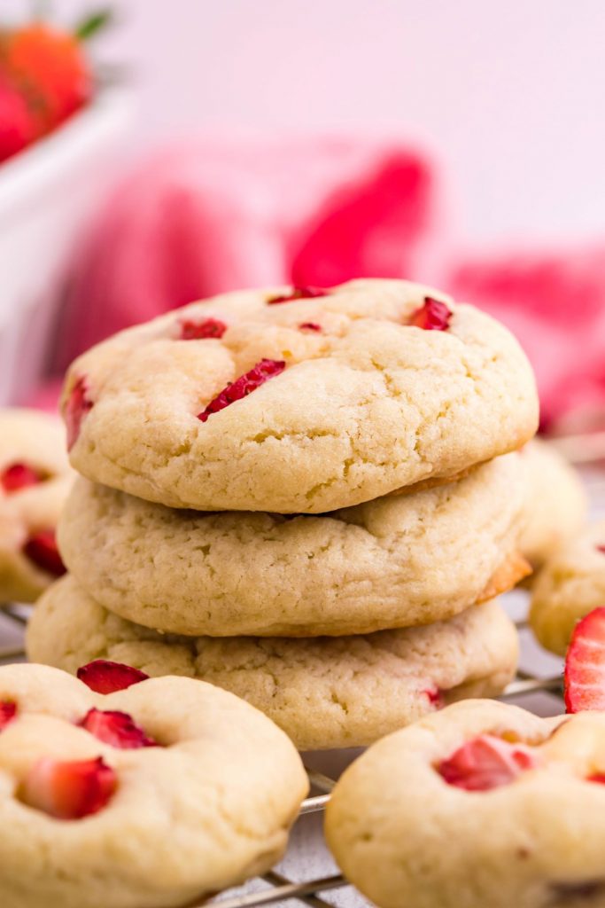 Three strawberry cheesecake cookies stacked on a gold wire rack with red linen. 