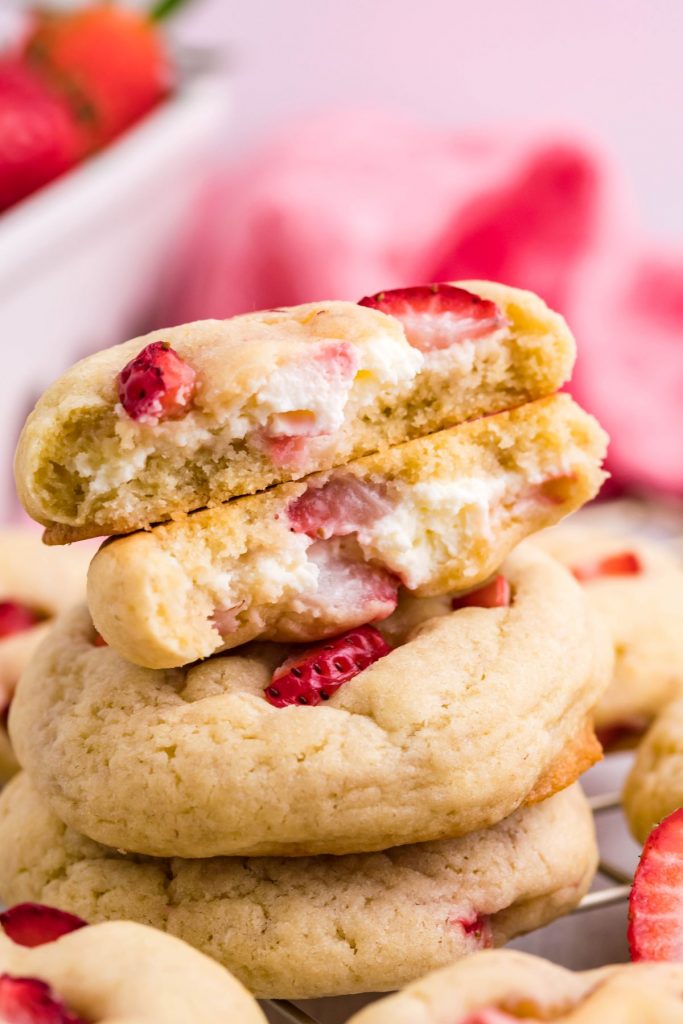 Three strawberry cheesecake cookies cut in half and stacked on a gold wire rack. Fresh strawberries peaking in from the side.  
