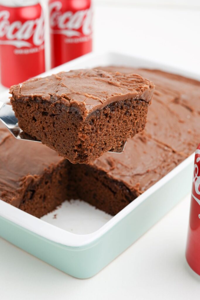A square piece of coca cola cake being removed from the baking dish being held with a silver spatula. Coke cans in he background. 