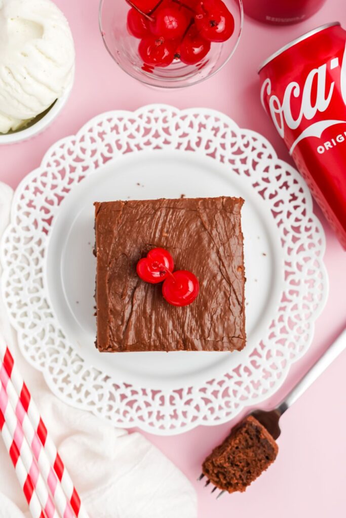 A square piece of coca cola cake on a white plate with two maraschino cherries on top. A can of coca cola peaking in on the side on a pink background. 