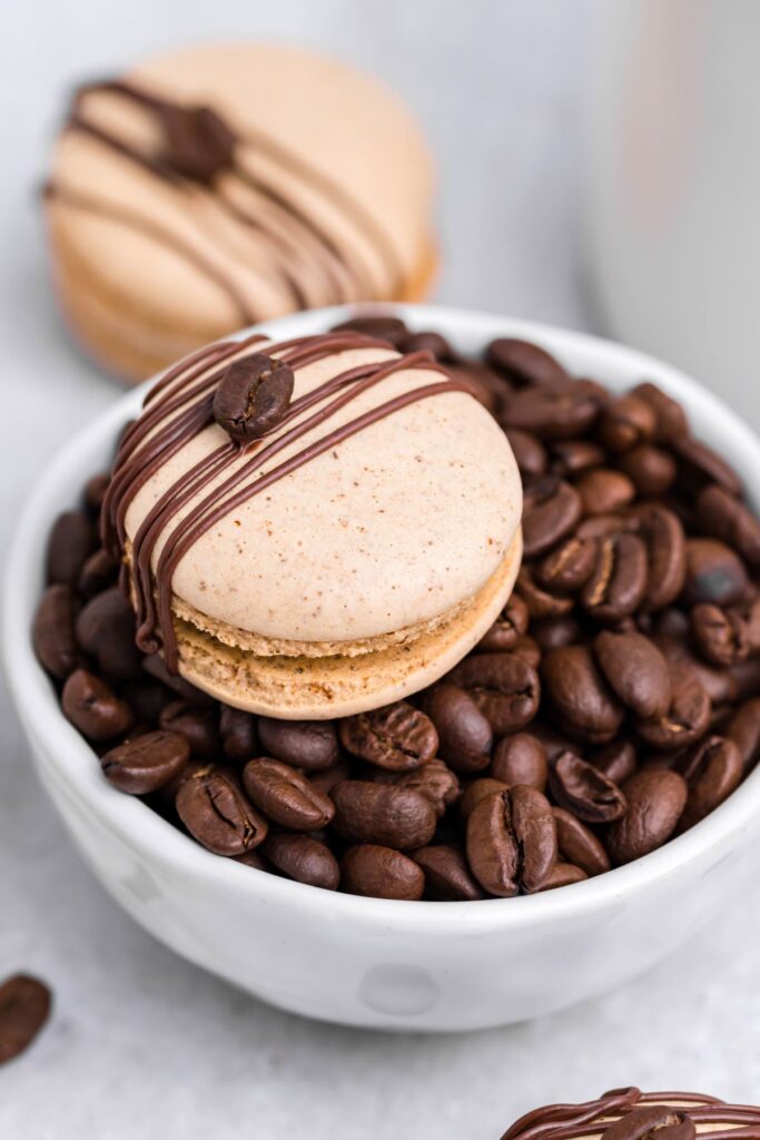 A coffee macaron sitting on a white bowl full of coffee beans. 