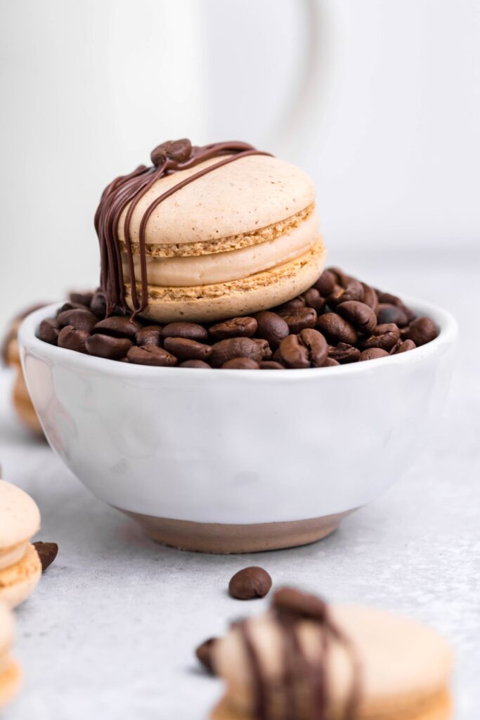 A coffee macaron sitting on a white bowl full of coffee beans. 