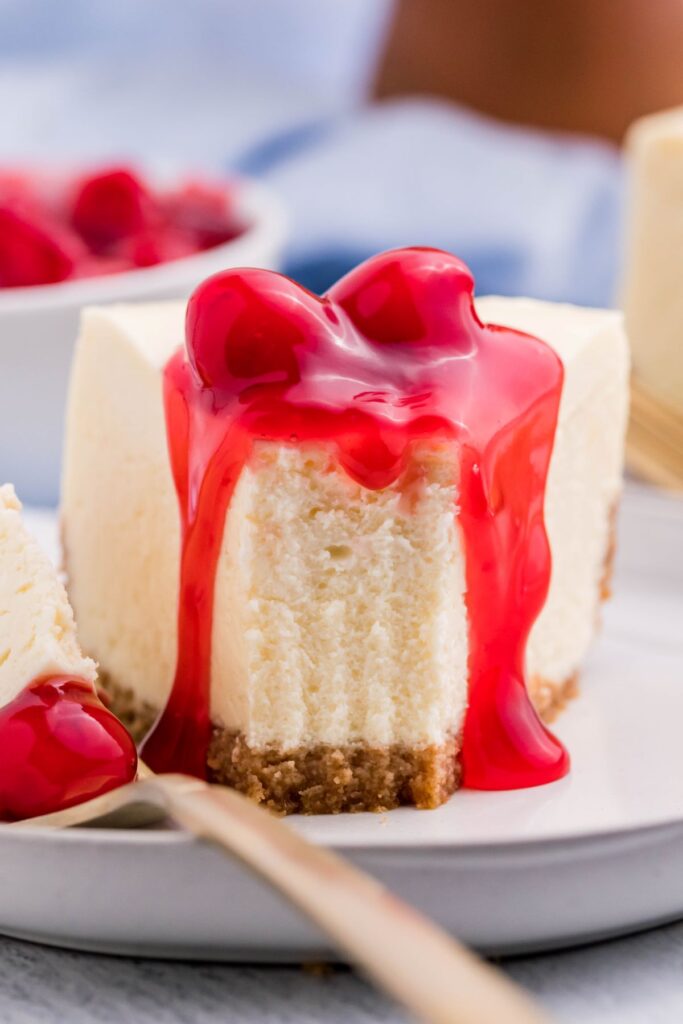 A front view of a piece of new york cheesecake with a bite removed on a gold fork with cherry topping on top. 