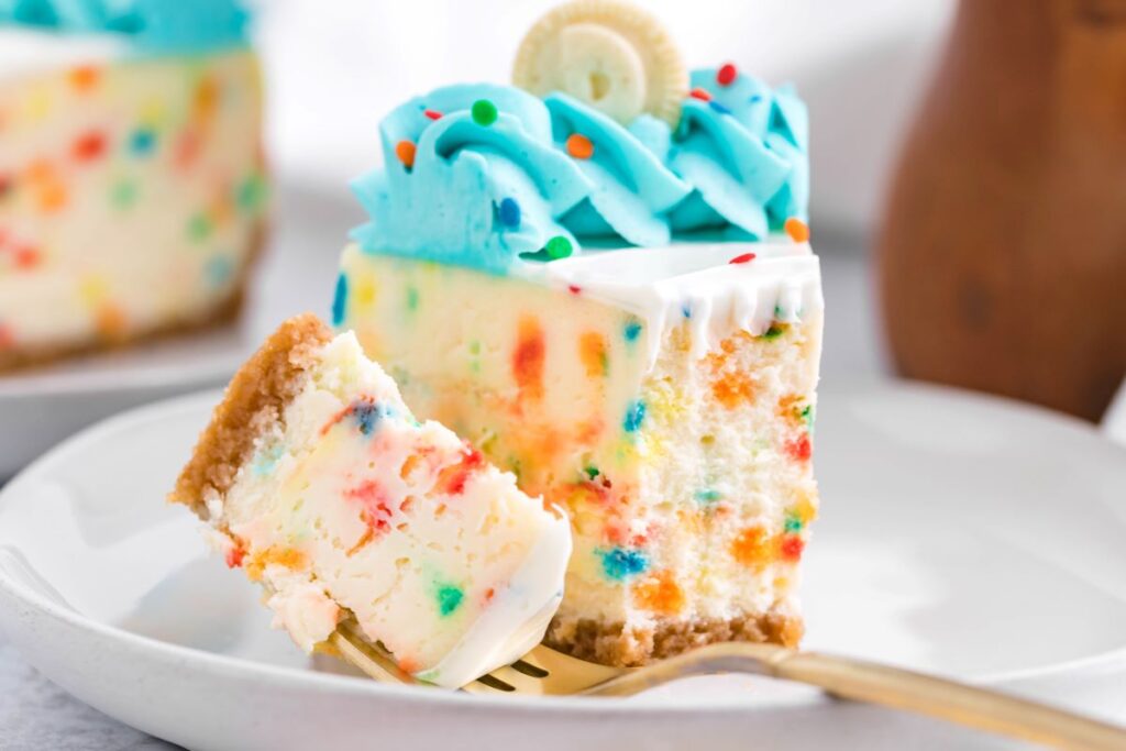 A piece of funfetti cheesecake on a white plate with a bite removed onto a gold fork. 