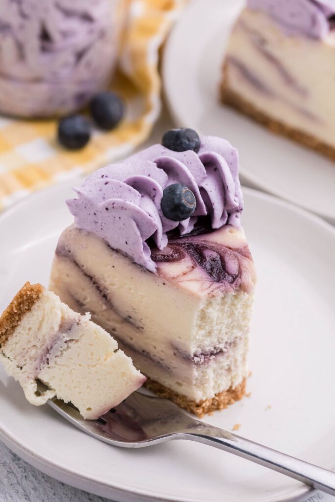  A slice of Instant pot blueberry swirl cheesecake on a white plate with a bite removed on a silver fork with yellow and white checkered linen in the background. 