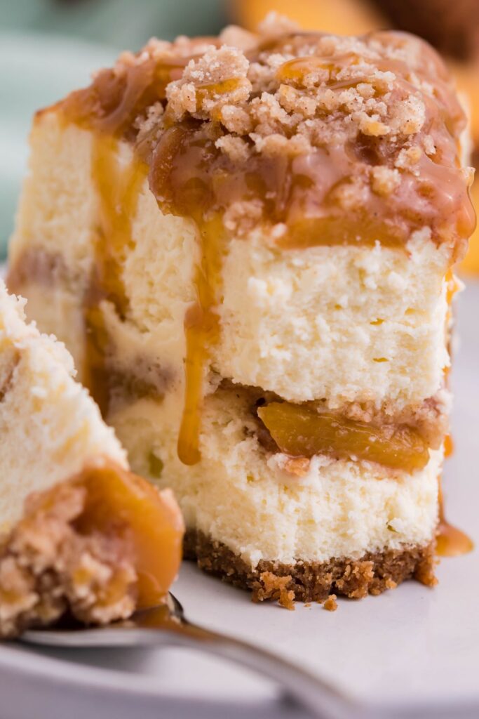 A piece of Instant Pot Peach Cobbler Cheesecake on a white plate with caramel drizzle with a bite removed to show the inside peach layer. 