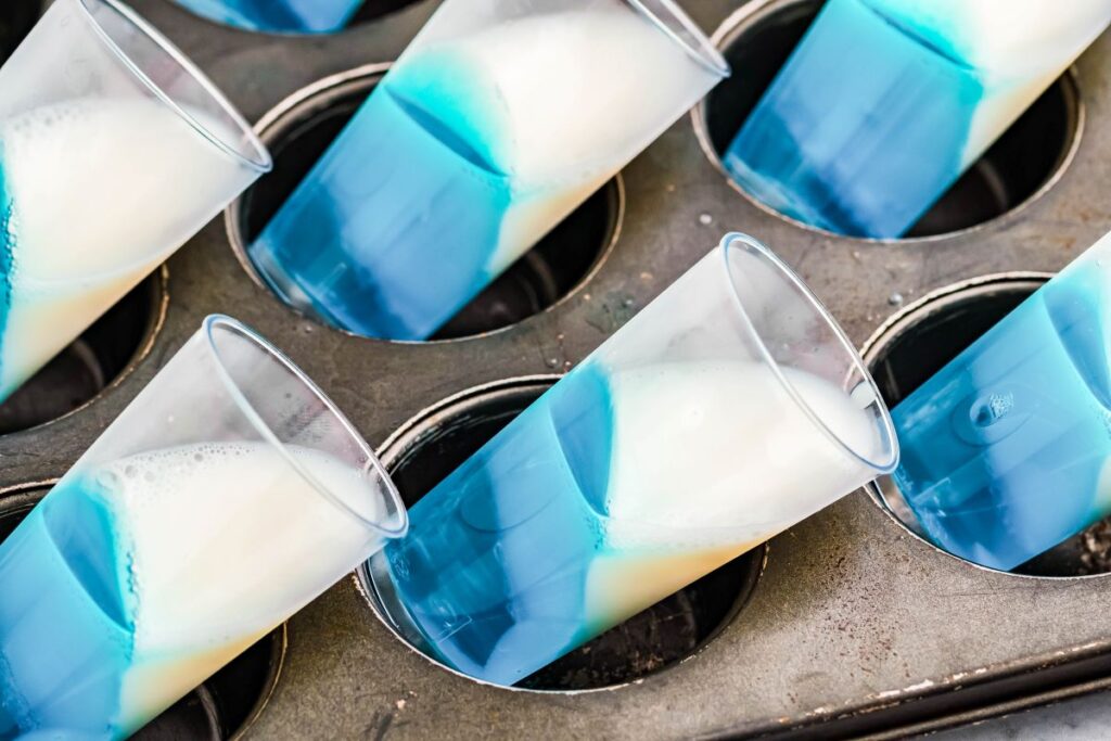 Tall shot glasses on their side in a muffin tin with blue and white jello layer. 