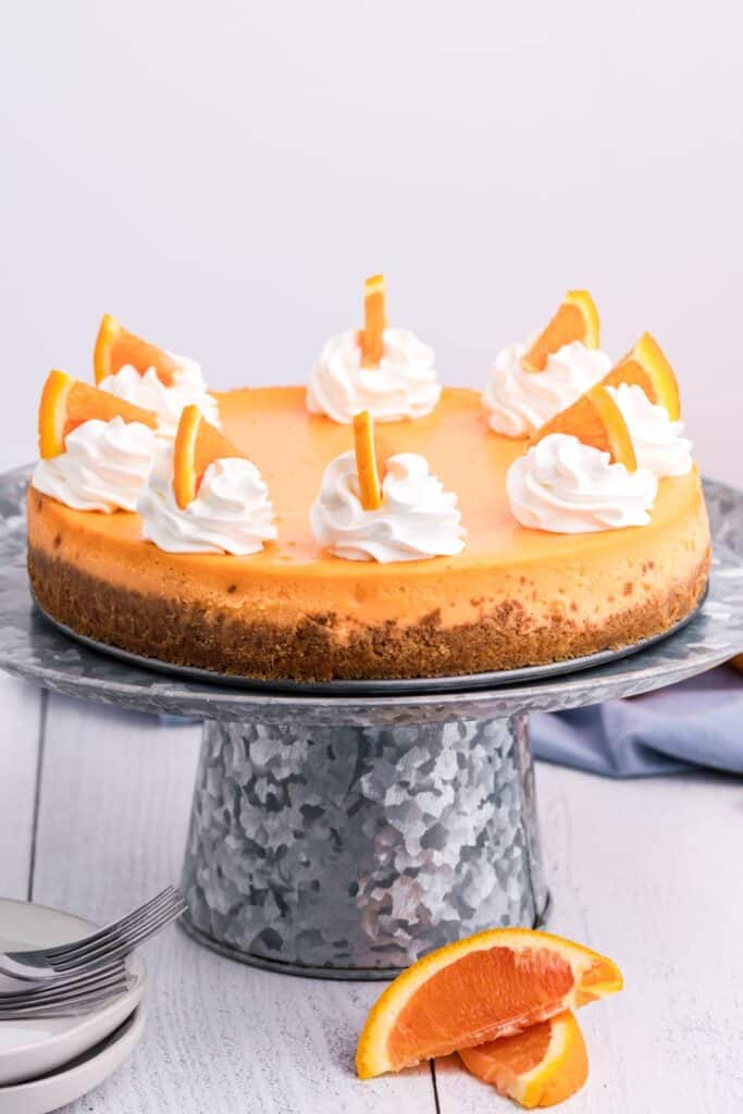 Orange Creamsicle Cheesecake on a tin cake treat with a blue linen, whipped cream swirls and oranges,