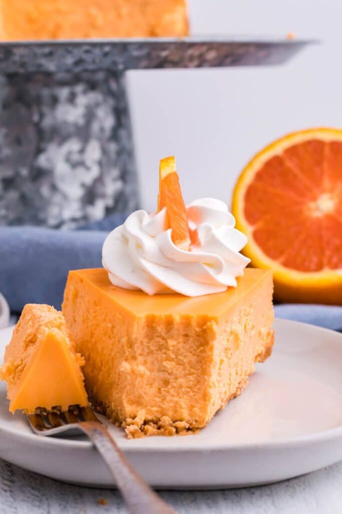 A slice of cheesecake on a white plate with a silver fork and blue linens and an orange in the background.