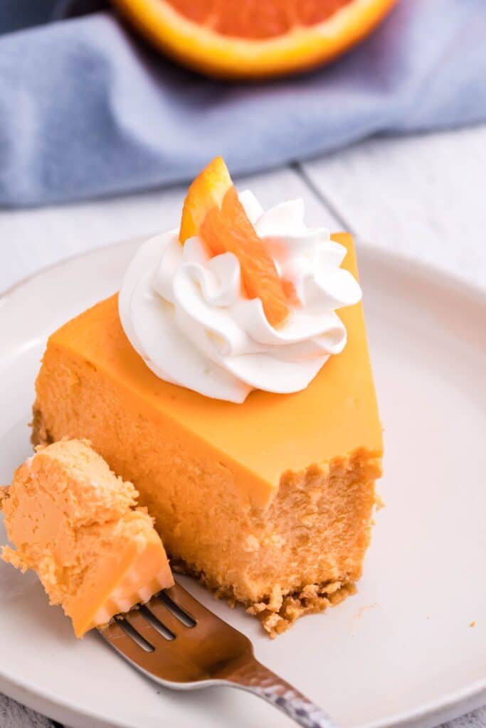 A slice of cheesecake on a white plate with a silver fork and blue linens and an orange in the background.
