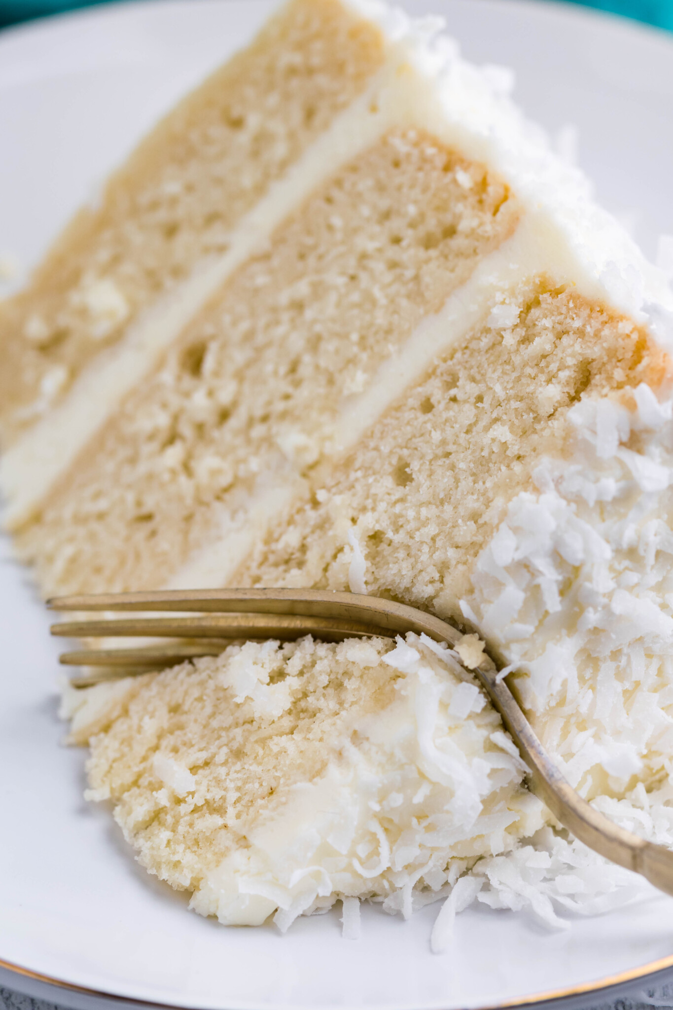 Close up view of a slice of Coconut Cake on a white plate served with a gold fork removing a bite.