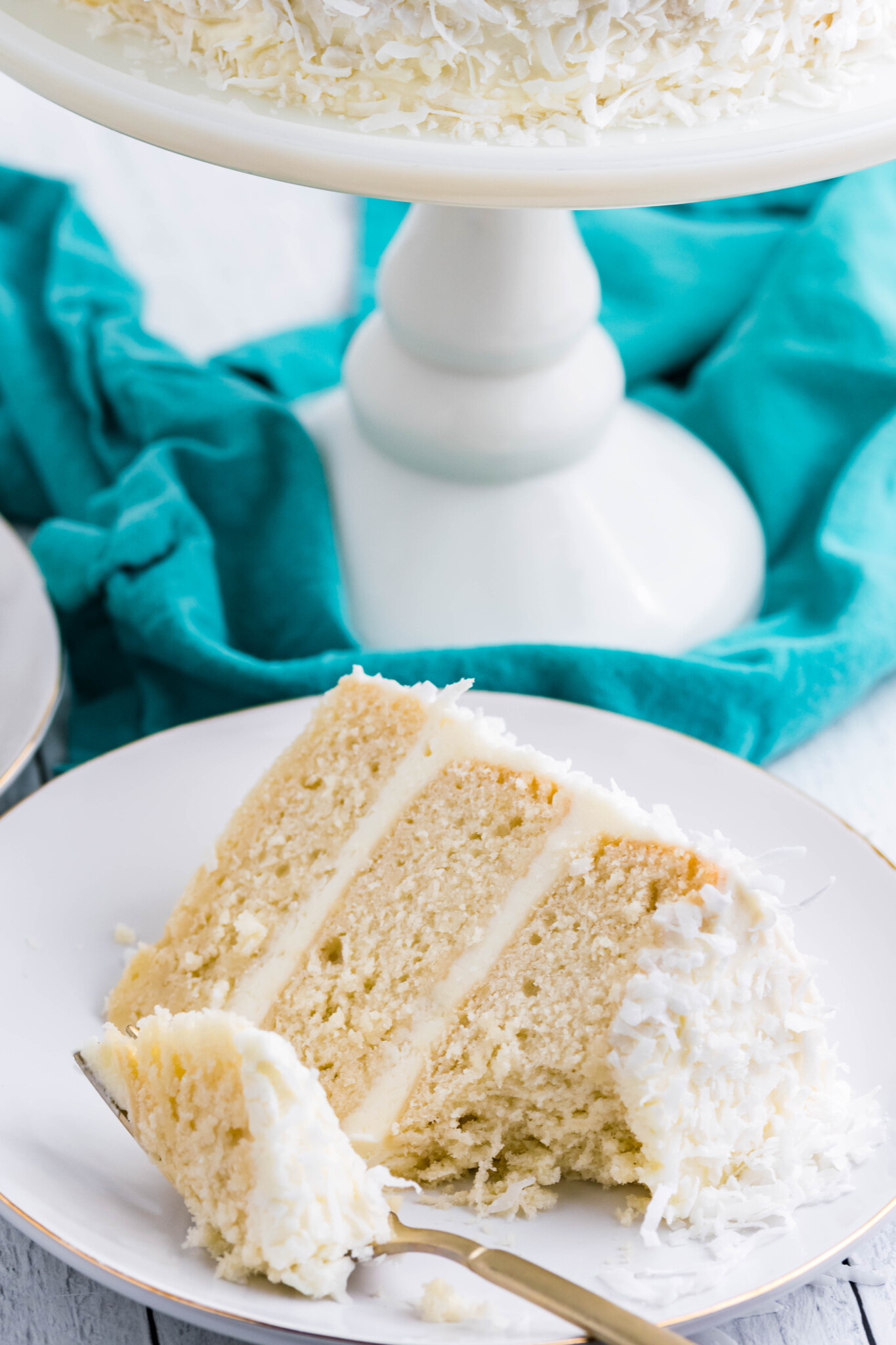 Front view of a slice of Coconut Cake on a white plate served with a bite removed by a gold fork.